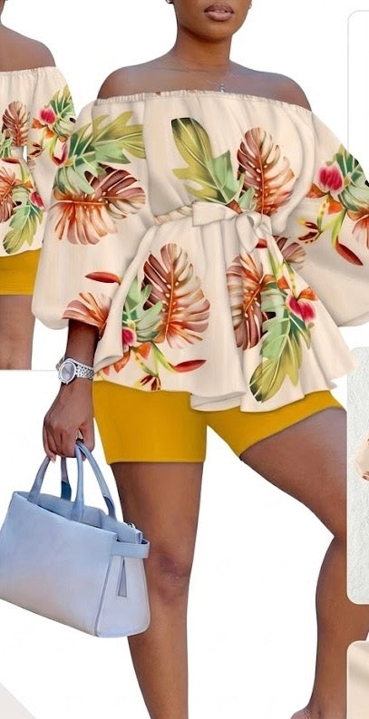 The Golden Hour Two-Piece Set is the calm confidence of a perfect moment — bottled in fabric. A warm champagne base blooms with coral, green, and red tropical florals across an off-shoulder peplum top with soft balloon sleeves — cinched with a matching belt tie and paired with rich gold biker shorts that glow against every skin tone.