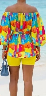 The Palette Two-Piece Set is for the woman who lives in full color. A vibrant abstract paint-block print in yellow, orange, teal, pink, and purple cascades across an off-shoulder peplum top with voluminous balloon sleeves — cinched at the waist with a matching belt tie and anchored by bold yellow biker shorts that pull the whole canvas together.