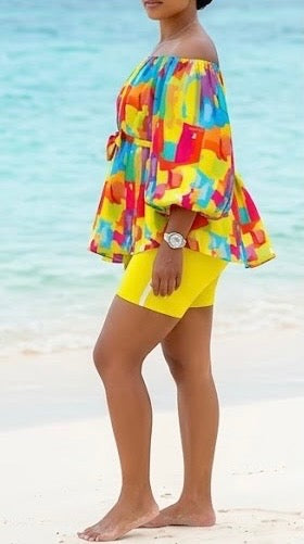 The Palette Two-Piece Set is for the woman who lives in full color. A vibrant abstract paint-block print in yellow, orange, teal, pink, and purple cascades across an off-shoulder peplum top with voluminous balloon sleeves — cinched at the waist with a matching belt tie and anchored by bold yellow biker shorts that pull the whole canvas together.
